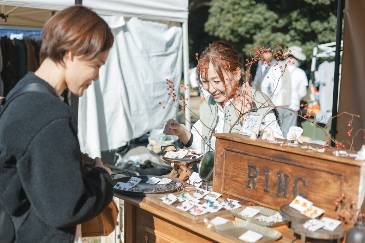 アクセサリーが並ぶグッズのブースで、商品を見ながら会話する出店者と来場者